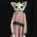 Die Katzen-Handpuppe, weisse Katze mit gestecktem Blümchen zwischen den rosa Ohren und in einem rosafarbenen Seidenkleid mit Stickereien und durchsichtigen Tüllärmeln.