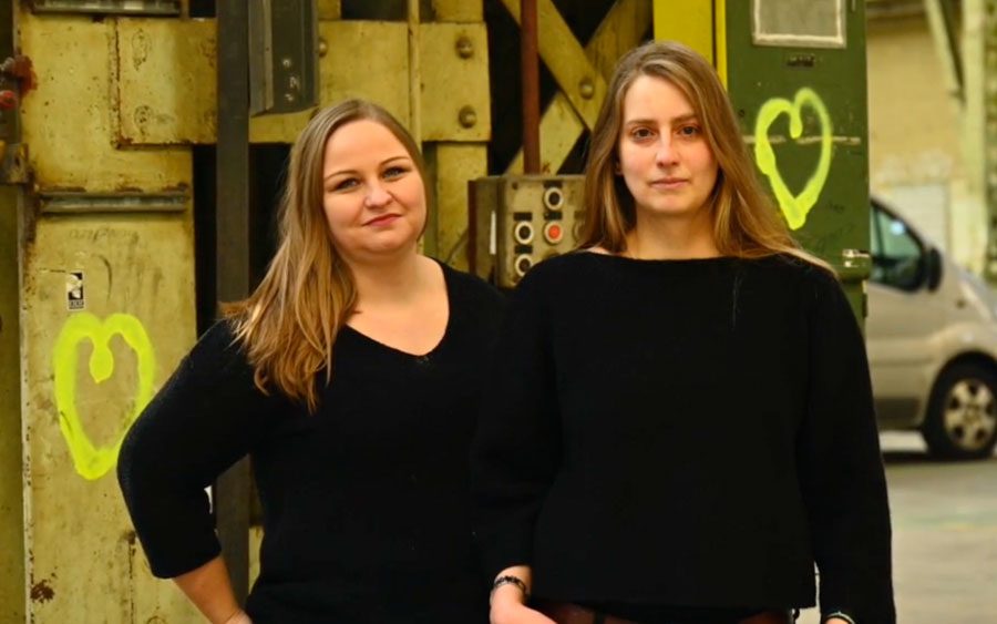 Zwei junge Theaterschaffende Frauen stehen in einer alten Fabrikhalle und schauen in die Kamera.
